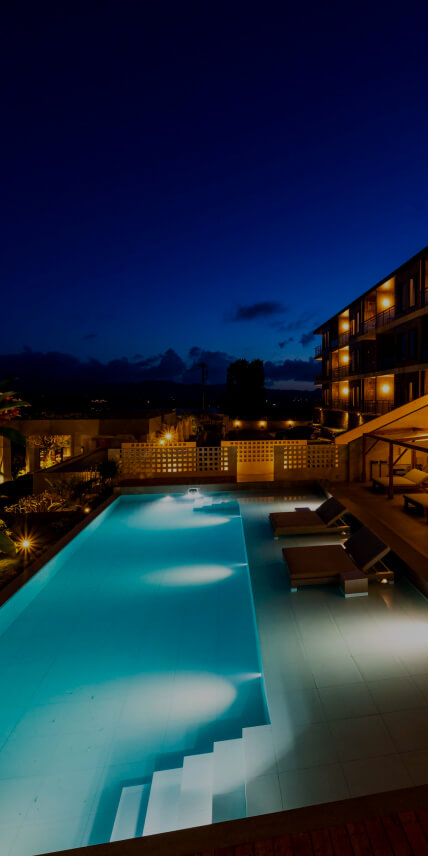 夜ライトアップされたプールサイド。広いプールの側にはゆっくり休めるソファや後ろにはホテルがある。