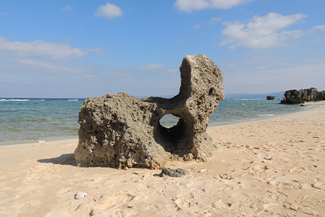 トケイ浜の白い砂浜に立つ、円形の穴が開いた独特な形状の岩(ポットホール)。岩の向こうには青く澄んだ海が広がり、遠くにはサンゴ礁の岩場が見える。空は青く、穏やかな波が打ち寄せる静かなビーチの風景。