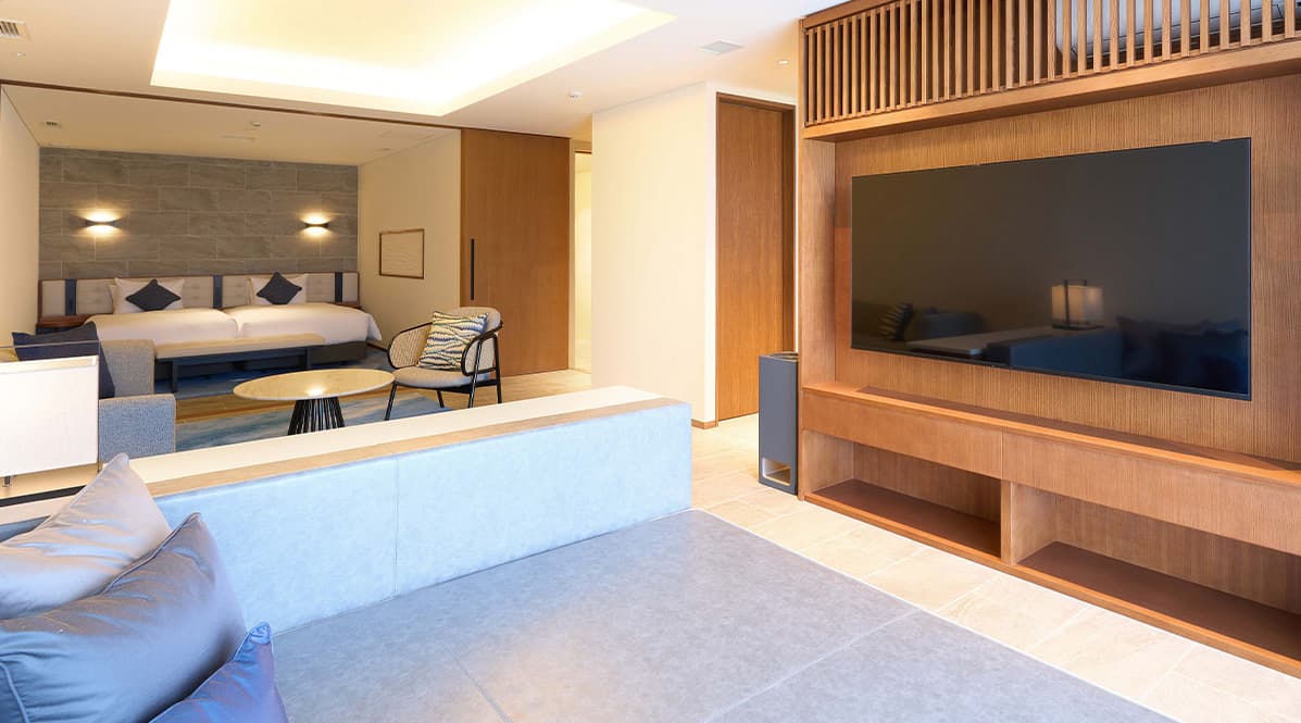 低めのソファと大きなテレビが備え付けられた木目調の収納があり、奥にはツインベッドが配置されたベッドルームが続きます。