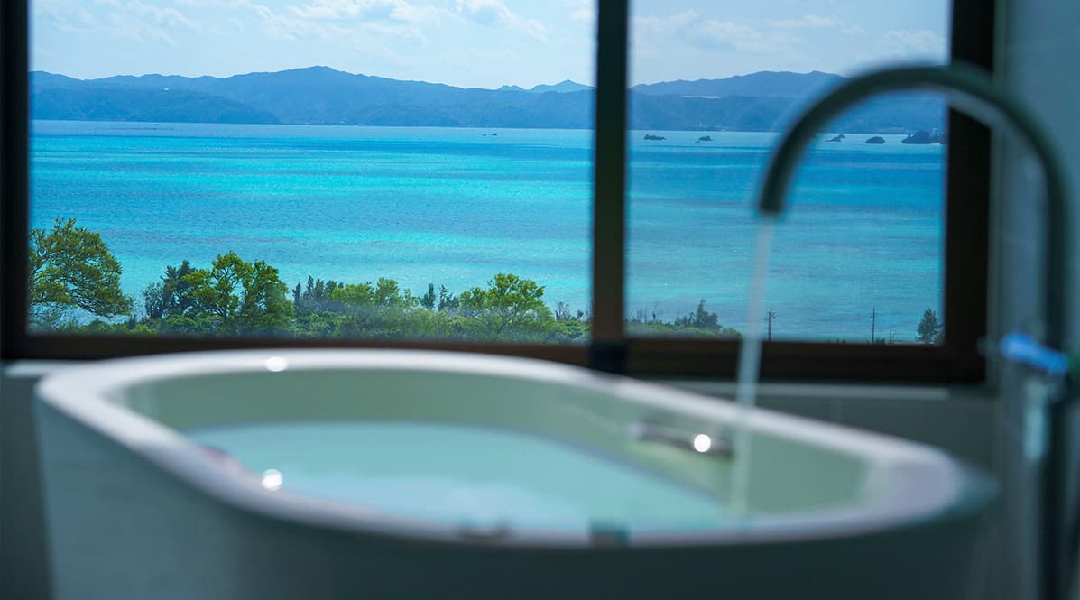 窓の外にエメラルドグリーンの海と遠くの山々が広がるオーシャンビューのバスルーム。白い湯船にはお湯が張られ、蛇口からは水が流れ出ています。