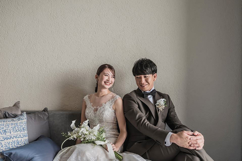 新郎新婦がグレーのソファに座ってくつろいでいる。新婦はオフホワイトのウェディングドレスを着用し、白いブーケを抱え、隣の新郎を見て明るく笑っている。新郎はダークブラウンのタキシードを着て、微笑みながら視線を新婦に向けている。