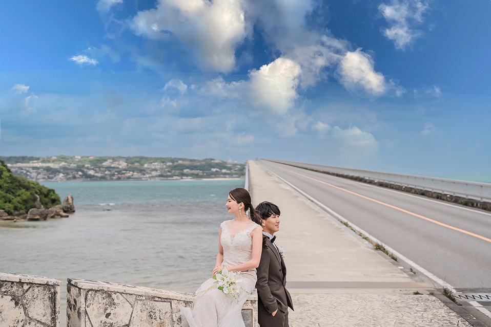 新郎新婦が沖縄の古宇利大橋の上で、海を背景に背中合わせに立っている。新婦はウェディングドレス姿で橋の欄干に座り、白いブーケを手に遠くを見ている。新郎はダークブラウンのタキシードを着用し、新婦に寄り添うように立って、カメラに視線を向けている。