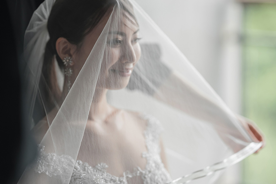 新婦がウェディングベールを顔の前に広げ、その薄い布越しに柔らかな笑顔を浮かべている。