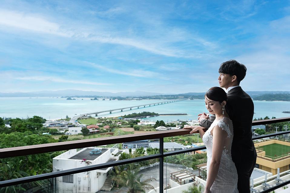 新郎新婦がホテルのバルコニーのような場所から、広大な沖縄の海と古宇利大橋の絶景を眺めている。新婦はオフホワイトのウェディングドレスを着用し、手すりに手を置いて横顔を見せている。新郎は黒いタキシード姿で、新婦の背後に寄り添うように立ち、同じく景色を見つめている。