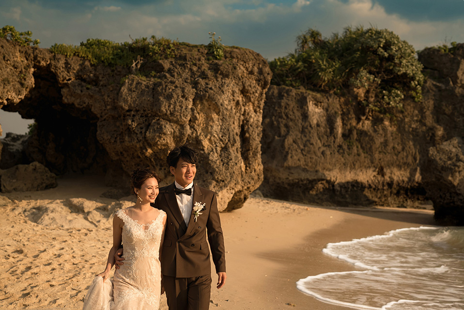 新郎新婦が沖縄のビーチで、夕暮れ時の柔らかな日差しの中、肩を並べて歩いている。新婦はオフホワイトのウェディングドレスを着用し、新郎の腕に軽く手を添え、優しく微笑んでいる。