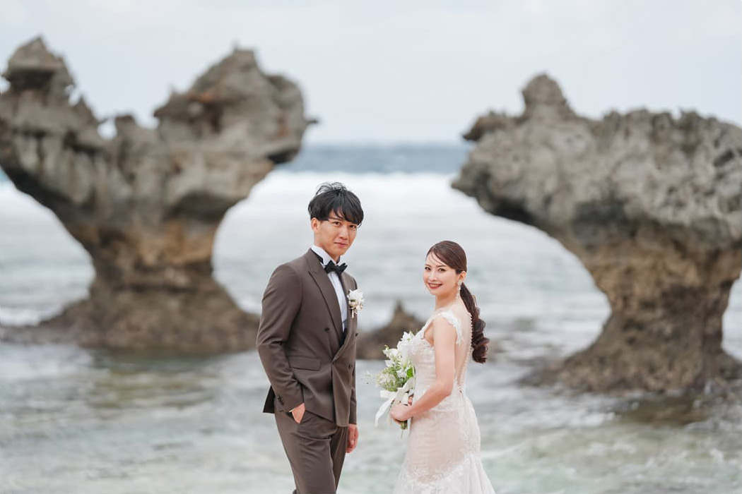 新郎新婦が沖縄のティーヌ浜にあるハートロックの前で、海を背景に立っている。新郎はダークブラウンのタキシードを着用し、ブートニアをつけている。新婦はオフホワイトのマーメイドラインのウェディングドレスを着て、白いブーケを持っている。