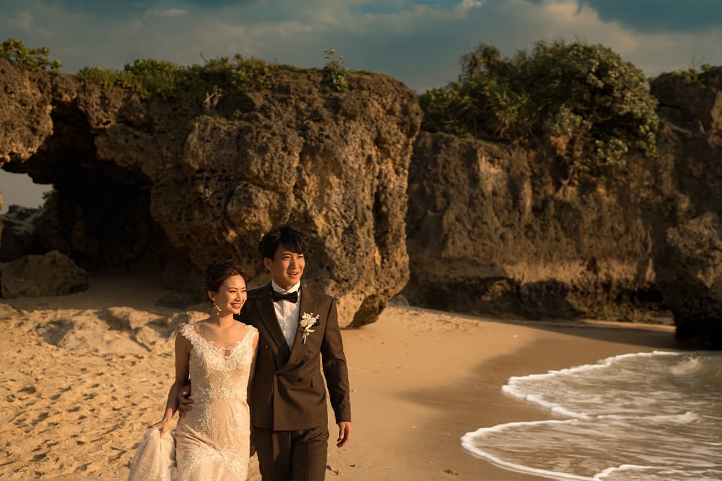 新郎新婦が沖縄のビーチで、夕暮れ時の柔らかな日差しの中、肩を並べて歩いている。新婦はオフホワイトのウェディングドレスを着用し、新郎の腕に軽く手を添え、優しく微笑んでいる。新郎はダークブラウンのタキシード姿で、前方を見つめている。背景には波打ち際と、特徴的な岩肌の洞窟や茂みが広がり、自然豊かな海岸の風景が写し出されている。