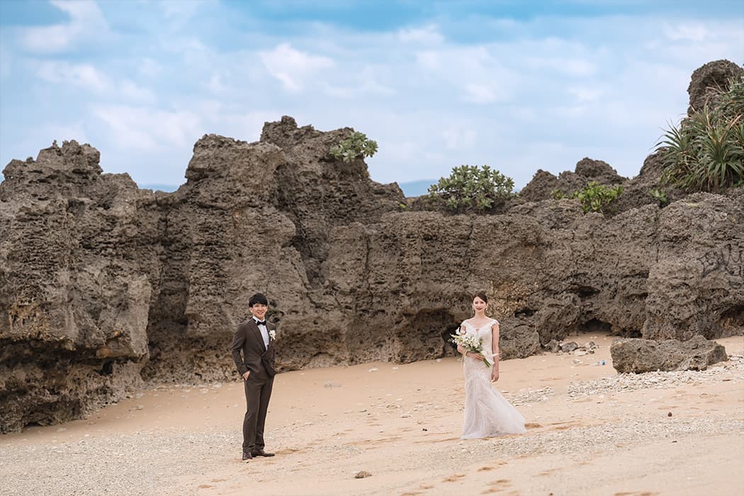 新郎新婦が沖縄のビーチで、背景に特徴的な岩が連なる場所で並んで立っている。新郎はダークブラウンのタキシードを着用し、カメラに向かって優しく微笑んでいる。新婦はオフホワイトのマーメイドラインのウェディングドレスを着て、白いブーケを手に、新郎と同じくカメラに視線を送っている。砂浜は広く、彼らの背後にはごつごつとした岩の崖が広がり、南国の植物が点々と生えている。