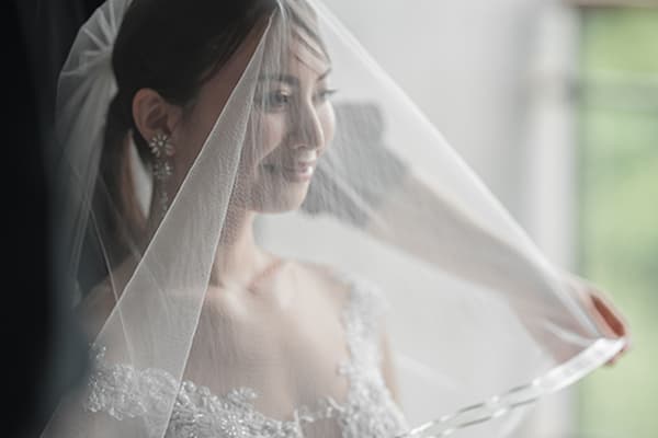 新婦がウェディングベールを顔の前に広げ、その薄い布越しに柔らかな笑顔を浮かべている。