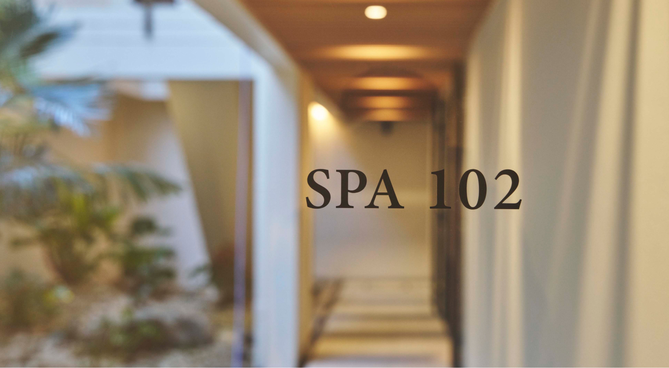 『SPA 102』と書かれたスパトリートメントルームの入口。廊下は木の天井と白い壁で構成され、間接照明で明るく照らされている。左側には緑豊かな植物が見える。